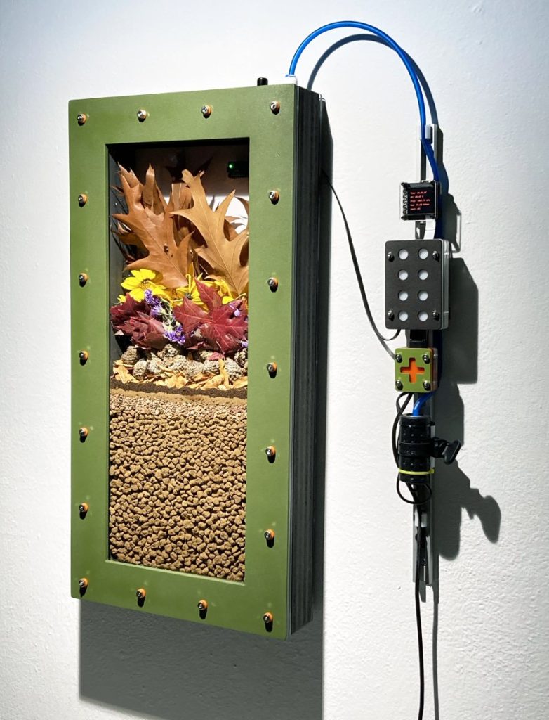 Moonwort Studio. Coulee Region -1983, 2023. Edaphotron: watertight enclosure, electronic gas sensor, microcontroller, bonsai gravel, plant parts, soil invertebrates and soil microflora. 15" X 4" X 23" (38cm X 8cm X 58cm).